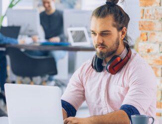 شاب مبدع يرتدي سماعات رأس يعمل على جهاز كمبيوتر محمول في بيئة مكتبية حديثة.